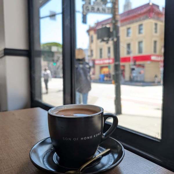 A "Son of Hong Kong" mug with a hot drink sits on a table by a window overlooking a city street.