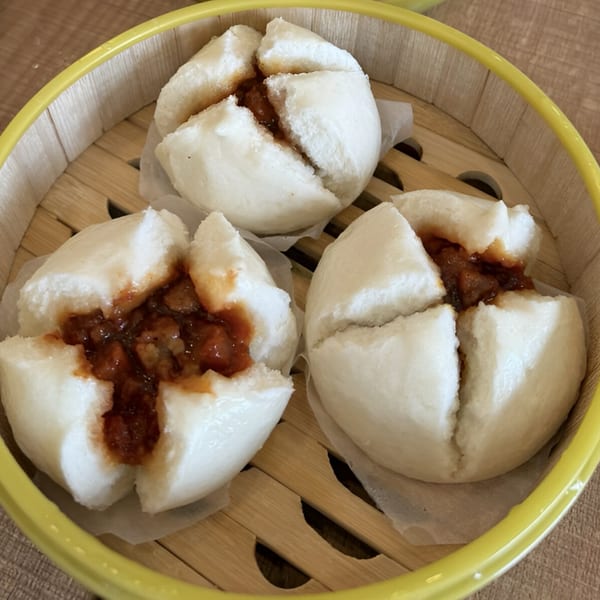 Three steamed char siu bao, white buns with savory pork filling, in a bamboo steamer.
