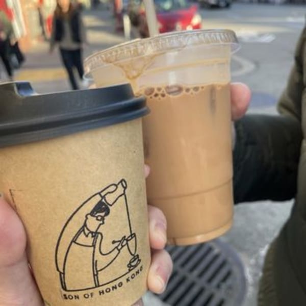 Two hands holding a paper cup and a plastic cup of iced coffee on a city street.
