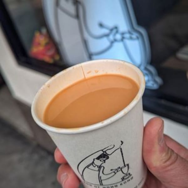 A hand holds a paper cup of Hong Kong-style milk tea with a cartoon drawing on the side.