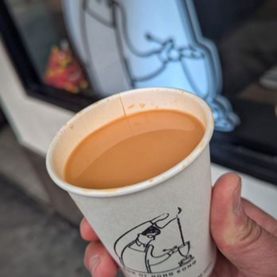 A hand holds a paper cup of Hong Kong-style milk tea with a cartoon drawing on the side.